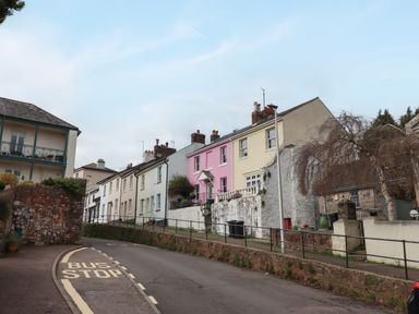 A street with pastel colored houses and a bus stop sign on the road at Rose Cottage in Bishopsteignton