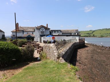 Outdoor seating area with wooden picnic tables next to a waterfront and a building in the background at Rose Cottage in Bishopsteignton