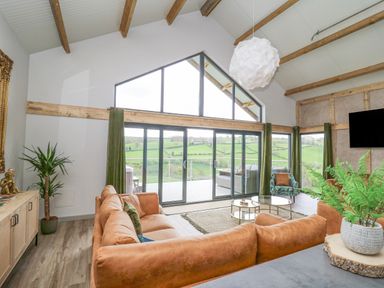 A living room with brown sofas a glass coffee table and large windows overlooking green fields at Barcud Coch - Red Kite in Garthmyl