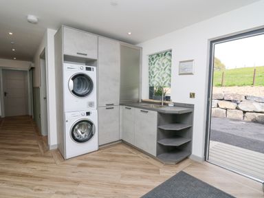 A utility room with a stacked washer and dryer unit next to cabinets a sink counters with shelves and a door leading outside at Barcud Coch - Red Kite in Garthmyl