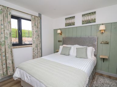 A bedroom with a bed with pillows and blankets a window with floral curtains and wall lights at Barcud Coch - Red Kite in Garthmyl
