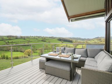 A patio with a glass table and cushioned wicker seats overlooking green rolling hills at Barcud Coch - Red Kite in Garthmyl