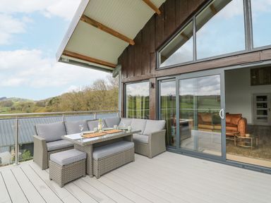 An outdoor patio with wicker seating and a glass table next to sliding glass doors at Barcud Coch - Red Kite in Garthmyl