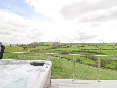 An outdoor hot tub on a deck overlooking green fields and hills at Barcud Coch - Red Kite in Garthmyl