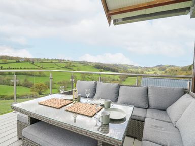 An outdoor seating area with a glass table set with cups plates and wine glasses overlooking green fields at Barcud Coch - Red Kite in Garthmyl