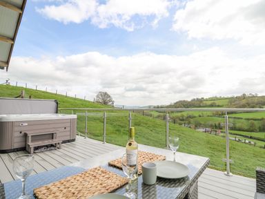 An outdoor deck with a hot tub a glass table set with wine glasses plates and a bottle overlooking green hills and fields at Barcud Coch - Red Kite in Garthmyl