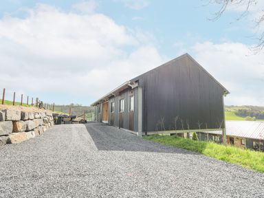 A gravel driveway leading to a black metal barn with windows and a stone retaining wall on the side at Barcud Coch - Red Kite in Garthmyl