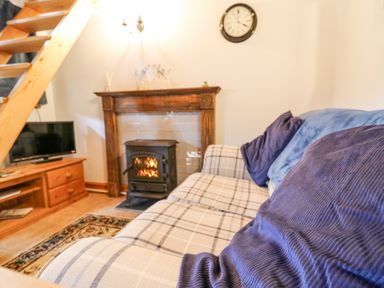 A living room with a sofa, cushions, a fireplace with a stove, a wooden TV stand, and a staircase at Ty Bach Twt in Synod Inn near New Quay