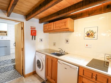 A kitchen area with wooden cabinets a washing machine a dishwasher a sink and tiled walls with fruit designs at Ty Bach Twt in Synod Inn near New Quay