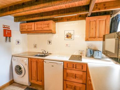 A kitchen with wooden cabinets a washing machine a fridge a sink a stovetop a kettle a toaster and a microwave at Ty Bach Twt in Synod Inn near New Quay
