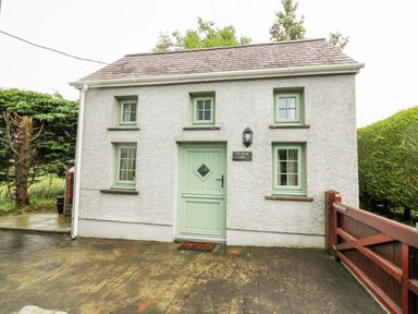 A small house with textured white walls green framed windows and door and a wooden gate at Ty Bach Twt Synod Inn near New Quay