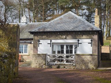 A small stone building with white wooden shutters and a gate surrounded by trees at Harness Lodge in Seahouses
