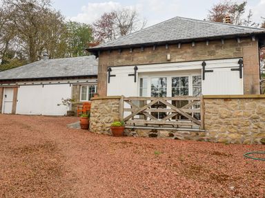 A stone building with white wooden doors and windows and a gravel driveway with potted plants at Harness Lodge in Seahouses