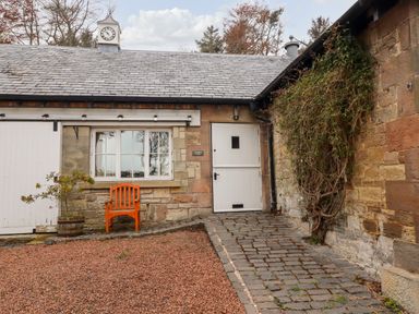 An exterior view of a stone building with a white door, a window, an orange wooden chair, and a small potted plant at Harness Lodge in Seahouses