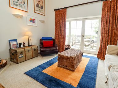 A living room with a wicker storage box on a blue and orange rug a leather armchair with red and blue cushions and a vintage trunk used as a table by the window at Harness Lodge in Seahouses