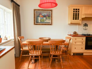 A kitchen dining area with a wooden table and four chairs a bowl on the table a wall painting a window with curtains and kitchen cabinets at Harness Lodge in Seahouses