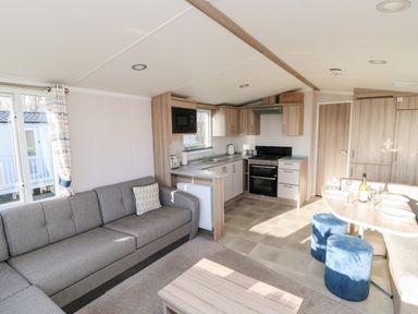 A living area with a gray sofa, wooden coffee table, kitchen with wooden cabinets, dining table with blue stools and set plates at 17 Summer Meadows in Ashington