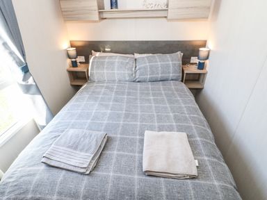 A bedroom with a double bed with gray checked bedding two folded towels on the bed bedside tables with lamps and a window with curtains at 17 Summer Meadows in Ashington