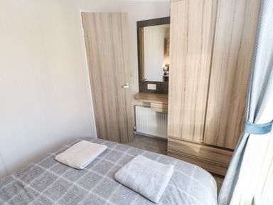 A bedroom with a bed, two folded towels, a wooden door, a wall mirror, a wooden wardrobe, and a curtain at 17 Summer Meadows in Ashington