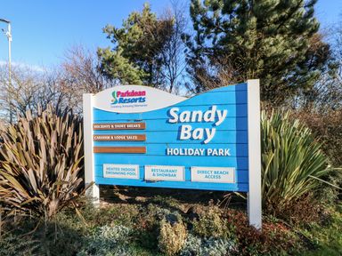 A blue sign for Sandy Bay Holiday Park with trees and plants around it at 17 Summer Meadows in Ashington