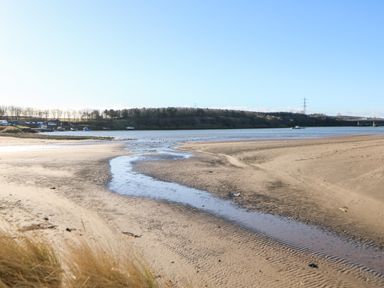 A sandy beach with a small stream leading to a body of water with boats and trees in the background at 17 Summer Meadows in Ashington
