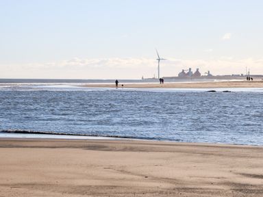 A beach with people walking near the water and an industrial area with a wind turbine in the distance at 17 Summer Meadows in Ashington