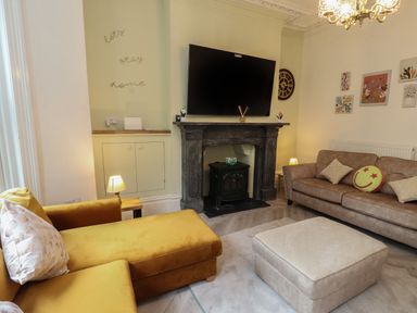 A living room with a yellow sofa, brown sofa, ottoman, TV above a fireplace, wall clock, and wall art at The Furness Haven in Barrow-In-Furness