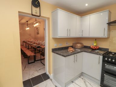 A kitchen with white cabinets and a countertop with bread and fruit next to a doorway leading to a dining area with a long table and benches at The Furness Haven in Barrow-In-Furness