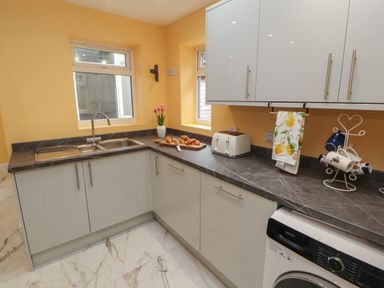 A kitchen with grey countertops white cabinets a toaster washing machine croissants on a cutting board and flowers in a vase at The Furness Haven in Barrow-In-Furness