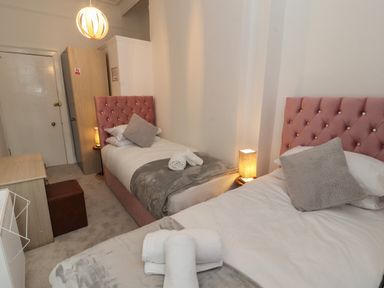 A bedroom with two beds with pink headboards towels on the beds bedside tables with lamps wardrobe and desk at The Furness Haven in Barrow-In-Furness