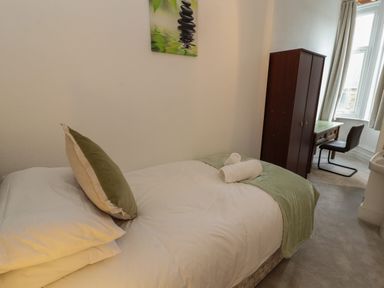 A bedroom with a single bed with pillows and a blanket a wooden wardrobe a desk with a chair and a window at The Furness Haven in Barrow-In-Furness