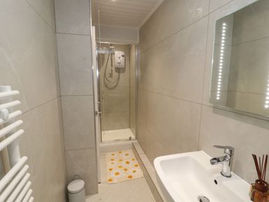 A narrow bathroom with a shower area a sink with faucet a wall mirror a white towel radiator and a trash bin at The Furness Haven in Barrow-In-Furness
