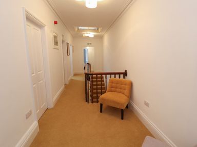 A hallway with beige carpet a wooden staircase and a mustard-colored chair at The Furness Haven in Barrow-In-Furness