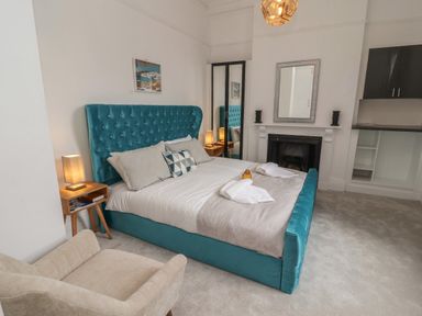 A bedroom with a turquoise upholstered bed a beige armchair bedside table lamps a fireplace and a mirror at The Furness Haven in Barrow-In-Furness