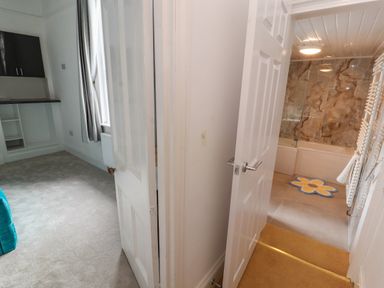A hallway with open doors leading to a carpeted room and a bathroom with a bathtub and floral rug at The Furness Haven in Barrow-In-Furness