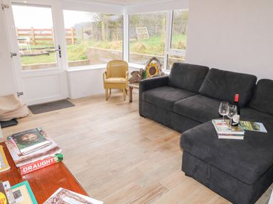 A sitting room with a black sofa a wooden chair a small table with wine bottle books and glasses and large windows overlooking a garden at The Poets Cottage in Portglenone