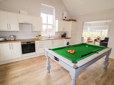 A kitchen area with white cabinets and wooden countertops next to a room with seating and a pool table at The Poets Cottage in Portglenone