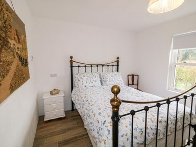 A bedroom with a metal bed frame with floral bedding a white bedside table with an object on it a wooden chair a window and a wall hanging at The Poets Cottage in Portglenone
