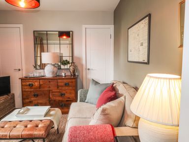 A living room with a beige sofa and colorful cushions a wooden chest of drawers with a lamp and decorative items a leather ottoman and a lamp at The Pottery in Newport Pembrokeshire