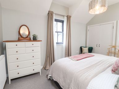 A bedroom with a white dresser with a mirror a window with beige curtains a bed with pillows and blanket and a wardrobe at The Pottery in Newport Pembrokeshire