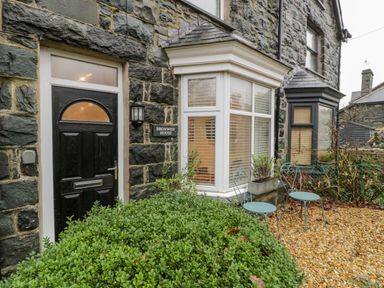 A stone exterior of a house with a black door a bay window and two metal chairs at Bronwen House in Llanbedr