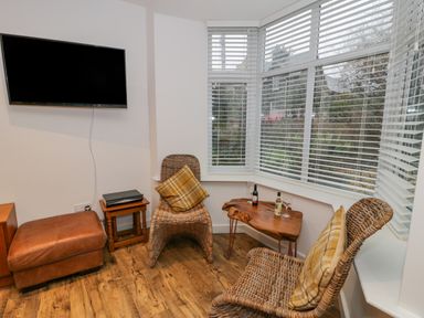 A corner seating area with two wicker chairs and plaid cushions a wooden table with bottles and glasses a wall-mounted television and a leather ottoman at Bronwen House in Llanbedr