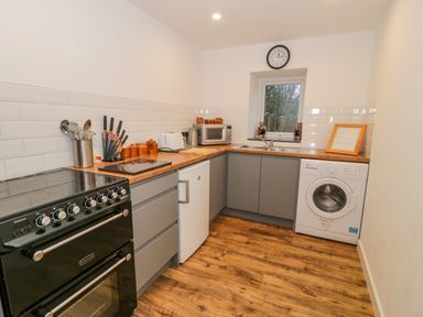 A kitchen with black stove, grey cabinets, wooden countertop, white fridge, microwave, toaster, kettle, washing machine, and a window at Bronwen House in Llanbedr