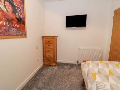 A bedroom corner with a wooden drawer chest a wall-mounted TV a radiator and a bed with a geometric patterned duvet at Bronwen House in Llanbedr