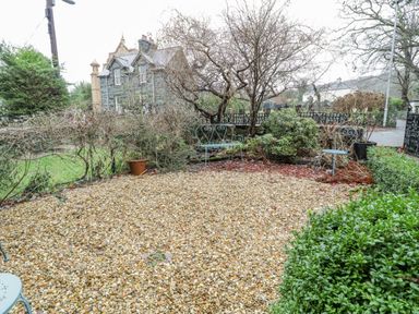 A small garden area with gravel ground, metal chairs, shrubs, and a stone house in the background at Bronwen House in Llanbedr