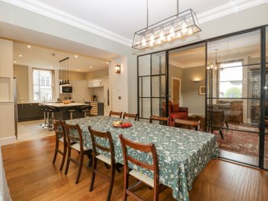A dining room with a long table covered by a patterned tablecloth and wooden chairs near a kitchen and a sitting area at Belle Vue in Hay on Wye Herefordshire