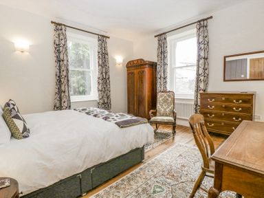 A bedroom with a bed a wooden wardrobe a chest of drawers an armchair a desk and two windows with curtains at Belle Vue in Hay on Wye Herefordshire