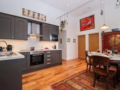 A kitchen with dark cabinets and stainless steel appliances next to a dining area with wooden chairs and a carpet at Ebenezer Chapel in Knodishall nr Saxmundham