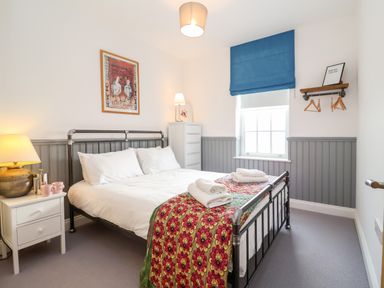 A bedroom with a double bed white bedding floral throw pillows bedside table with lamp and cups a window with blue blind and a wall shelf with hangers at Ebenezer Chapel in Knodishall nr Saxmundham