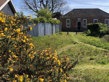 A grassy path leading to a brick house with a dark door and white windows behind a metal fence and flowering yellow bushes in a garden at Ebenezer Chapel in Knodishall nr Saxmundham